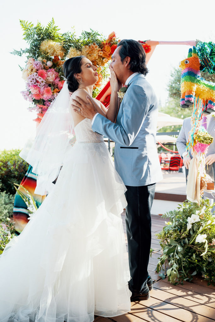 bride wipes tears from grooms face in front of colorful arch, mexican theme, barcelona brides